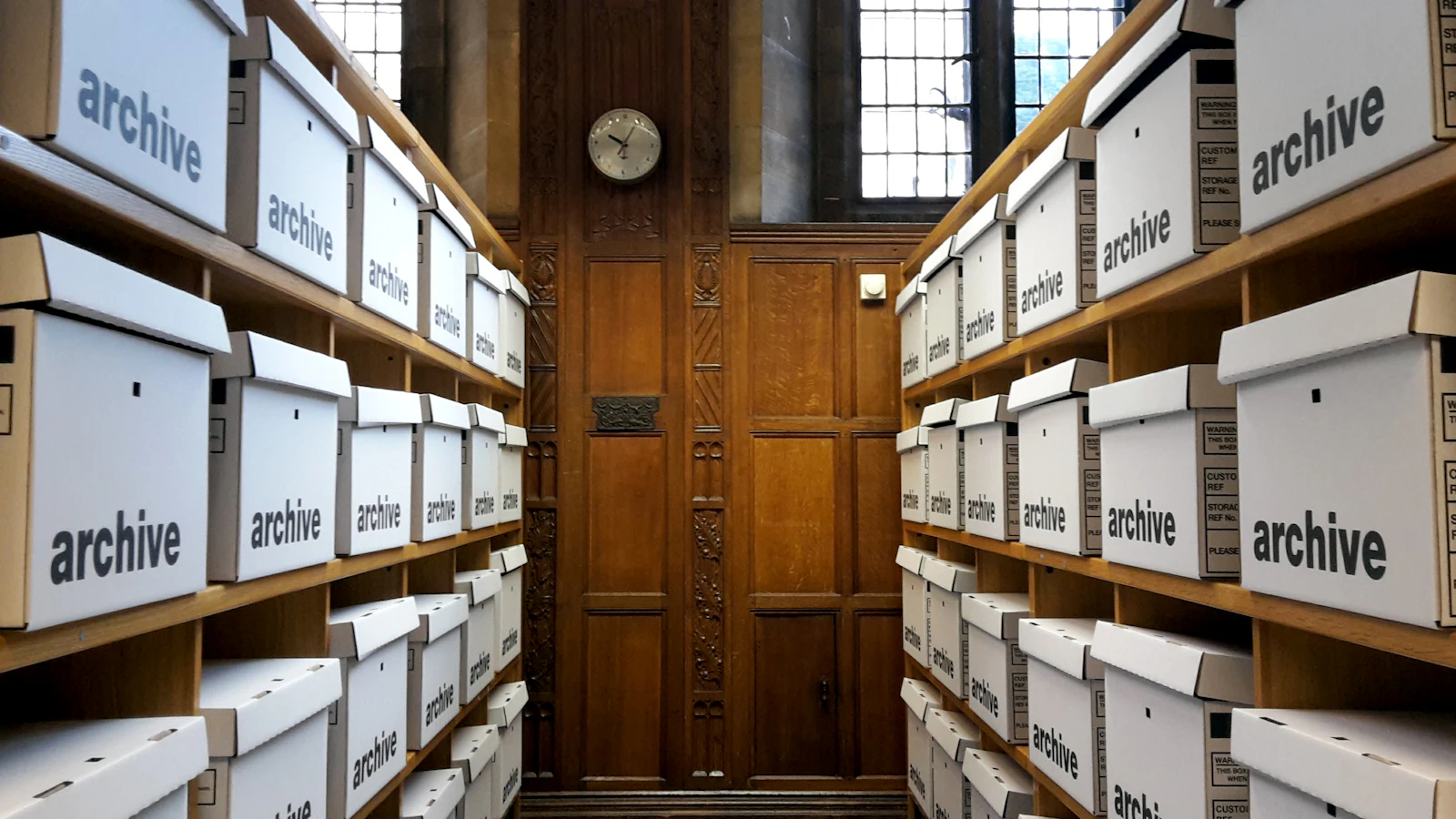 Regalgang in einem Aktenarchiv mit beschrifteten Archivkartons und einer Wanduhr an der Stirnwand