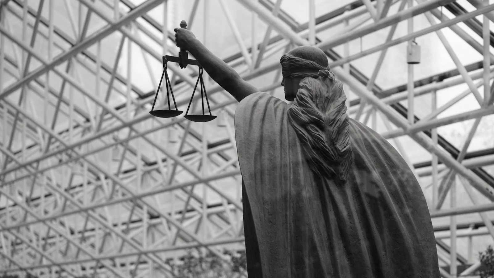 Schwarzweiße Justitia-Statue von hinten, mit ausgestreckter Hand und Waage, unter einem Glasdach